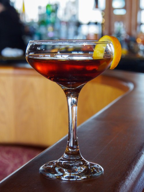 A stylish cocktail in a coupe glass, garnished with a twist of orange peel, sits on a wooden bar.