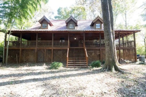 A wooden cabin with a red metal roof, featuring a spacious porch and surrounded by trees in a natural setting.