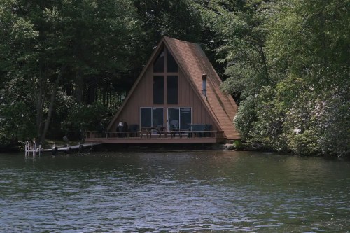 A lakeside A-frame cabin surrounded by trees, featuring a deck and large windows overlooking the water.
