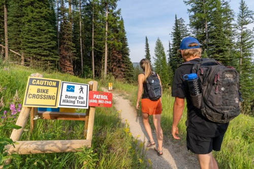 Two hikers walk along a trail surrounded by trees and wildflowers, with caution signs for bike crossings.