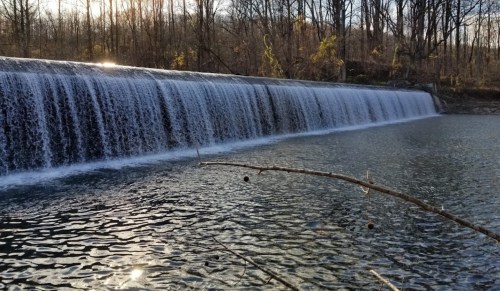 A serene waterfall cascading over a dam into a calm river, surrounded by bare trees and soft sunlight.
