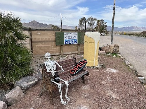 A skeleton sits on a bench next to a sign that says "OPEN" and a large beer mug, with desert scenery in the background.