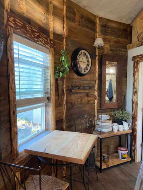 Cozy dining nook with a wooden table, chairs, wall clock, mirror, and plants in a rustic interior.