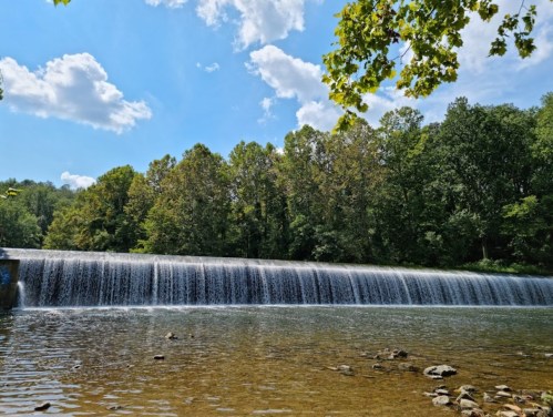 A serene waterfall cascades over a dam, surrounded by lush green trees and a clear blue sky with fluffy clouds.