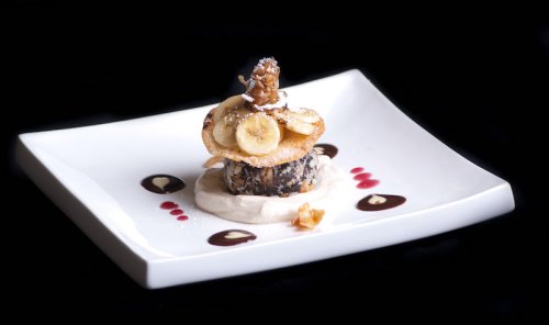 A gourmet dessert featuring layers of chocolate, banana slices, and a crispy topping, drizzled with sauce on a white plate.