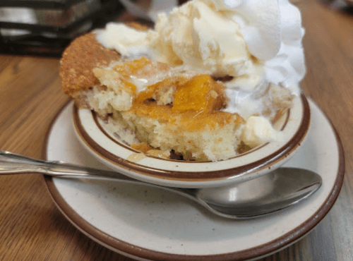 A slice of warm dessert topped with melting ice cream on a plate, with a spoon beside it.
