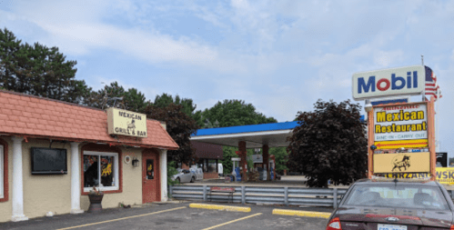 A Mexican restaurant with a grill and bar next to a Mobil gas station, surrounded by trees and a cloudy sky.