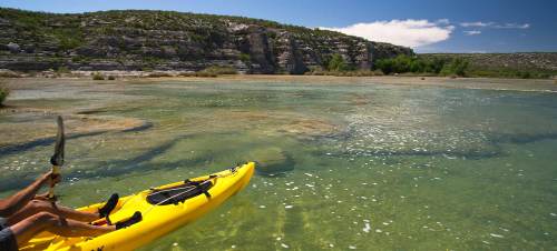 A person paddles a yellow kayak on a clear river surrounded by rocky cliffs and lush greenery under a blue sky.