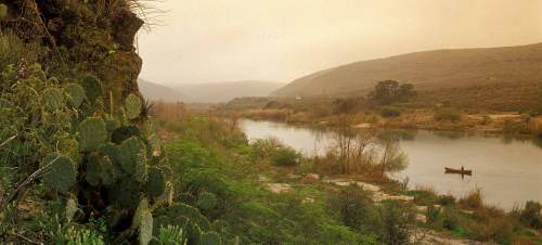 A serene river landscape with hills, cacti, and a small boat on the water under a hazy sky.