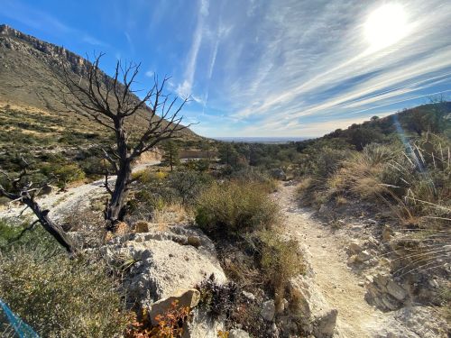 A scenic landscape featuring a winding trail, dry vegetation, and a bright sun against a blue sky with wispy clouds.