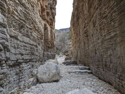 Narrow canyon with steep rock walls and a dry riverbed, featuring large boulders and sparse vegetation.
