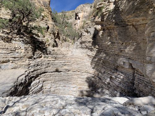 A rocky canyon with layered stone walls and sparse vegetation under a clear blue sky.