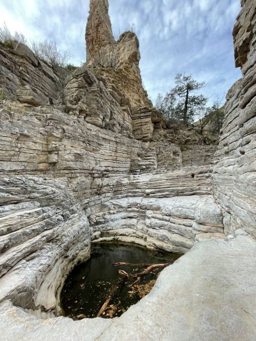 Rocky canyon with layered stone walls and a small pool of water at the bottom, under a cloudy sky.