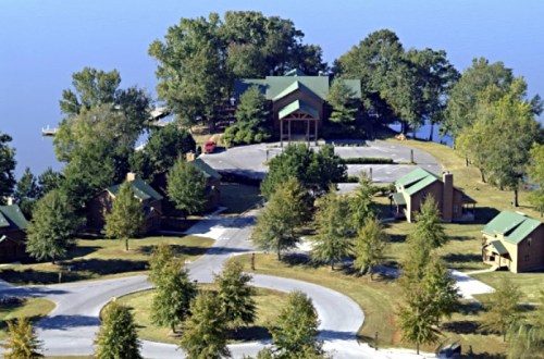 Aerial view of a lakeside lodge surrounded by trees and winding roads, with cabins and a parking area visible.