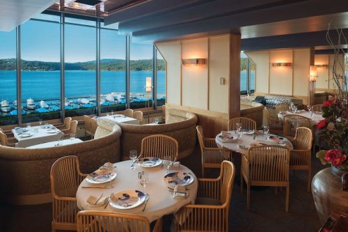 Elegant restaurant interior with round tables, overlooking a scenic lake and mountains through large windows.