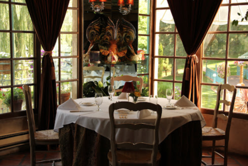 A cozy restaurant table set for two, with large windows showcasing a garden view and elegant decor.