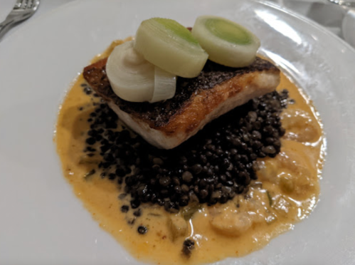 A plated dish featuring seared fish on a bed of black lentils, topped with sliced leeks and a creamy sauce.