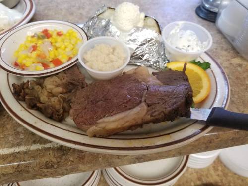 A plate with sliced roast beef, corn salad, baked potato, and sides of sour cream and butter, garnished with an orange slice.