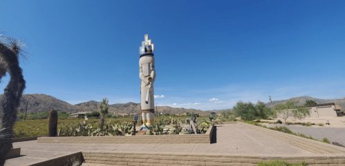 A tall, abstract sculpture stands in a desert landscape under a clear blue sky, surrounded by cacti and mountains.