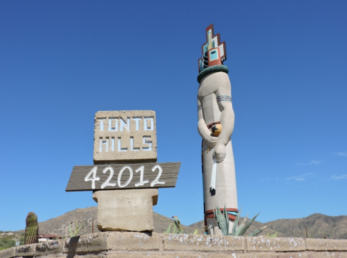 A large statue stands next to a sign reading "Tonto Hills 42012" against a clear blue sky and desert landscape.