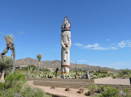 A tall, decorative totem stands in a desert landscape, surrounded by cacti and mountains under a clear blue sky.