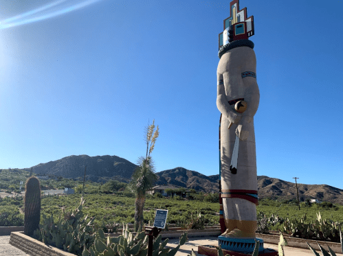 A large statue of a figure stands in a desert landscape with mountains in the background and cacti in the foreground.