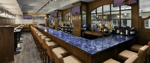 A modern bar with a long blue countertop, wooden accents, and shelves stocked with various bottles.