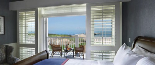 A cozy hotel room with a view of the ocean through open shutters and a small balcony with chairs.
