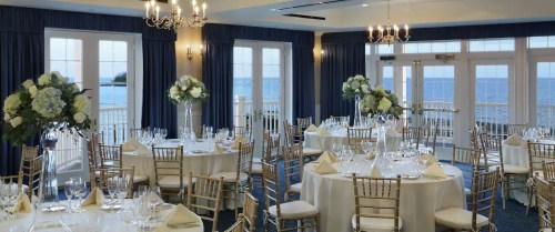 Elegant dining room with round tables, white linens, floral centerpieces, and a view of the ocean through large windows.