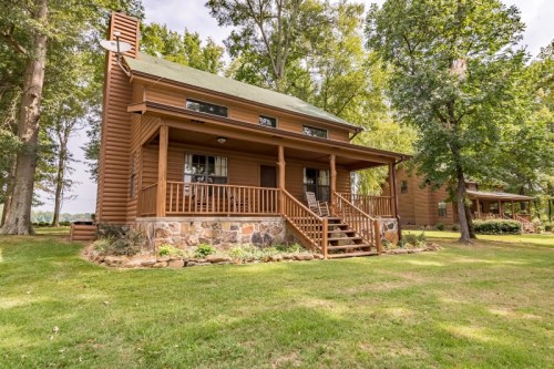 A two-story wooden cabin with a green roof, surrounded by trees and a grassy area. Steps lead to the front porch.