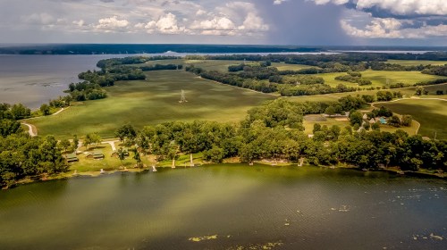 Aerial view of a lush green landscape with a lake, trees, and distant hills under a partly cloudy sky.
