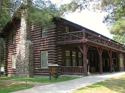 A rustic log cabin with a stone chimney, featuring a balcony and surrounded by trees and grass.