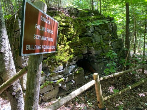 A moss-covered stone structure in a forest, with a warning sign about falling rocks nearby.