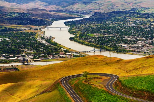 A winding road leads down a hillside overlooking a river and a town surrounded by rolling green hills.