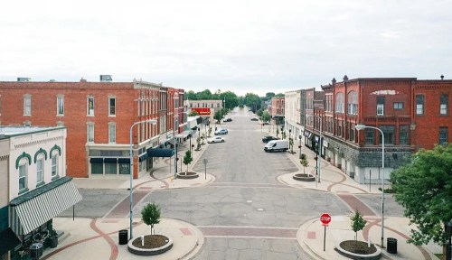 A quiet, empty street lined with brick buildings and trees, featuring parked cars and a stop sign.