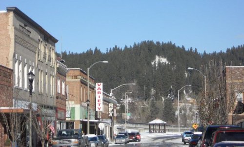 A quaint downtown street lined with shops, trees, and a backdrop of snow-capped hills under a clear blue sky.