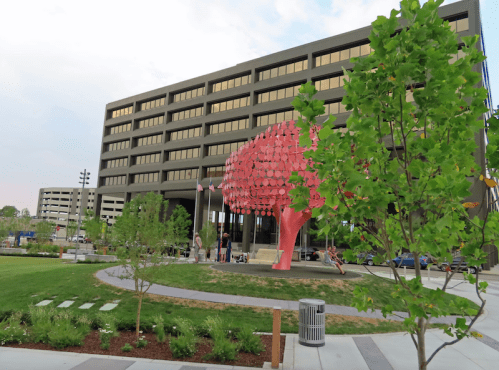 A modern building with large windows, featuring a vibrant red sculpture and landscaped greenery in the foreground.