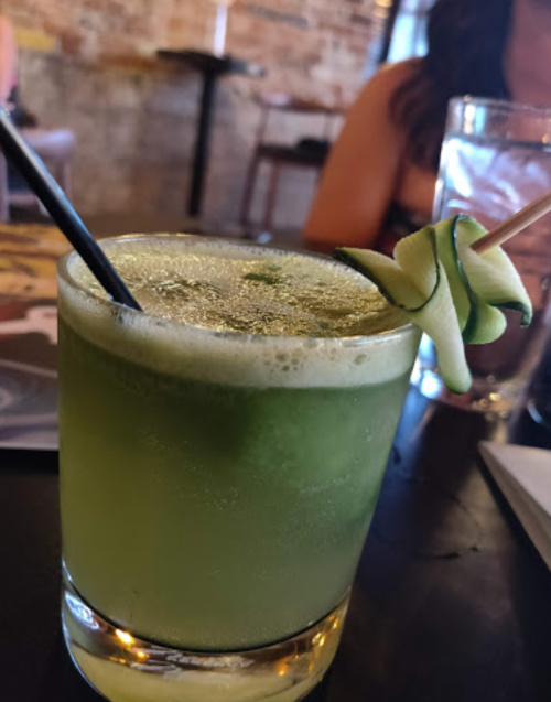A green cocktail garnished with a spiral cucumber slice, served in a glass with a straw on a dark table.