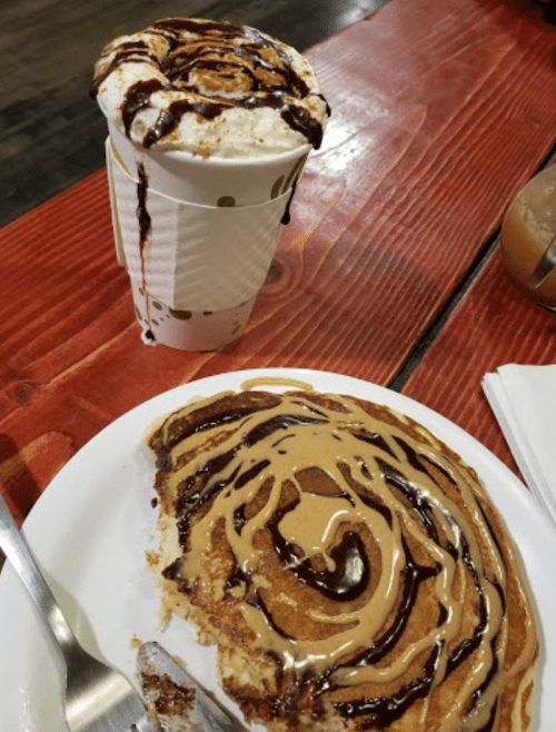 A plate with a cinnamon roll topped with chocolate and peanut butter, next to a cup of hot chocolate with chocolate drizzle.