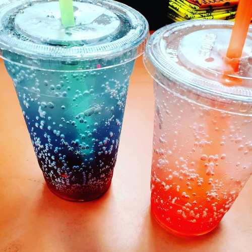 Two colorful drinks in clear cups with bubbles, one blue and one pink, placed on an orange surface.