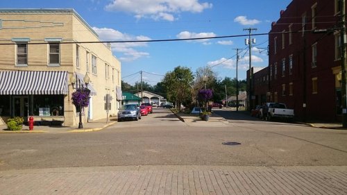 A small town street with buildings, parked cars, and hanging flower baskets under a blue sky with clouds.