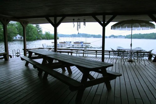 A covered wooden deck overlooking a lake, featuring picnic tables and a patio umbrella, with boats in the background.