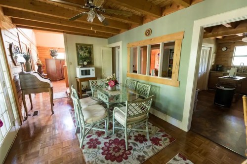 Cozy dining area with a glass table, chairs, and floral rug, leading to a kitchen and living space in a rustic home.