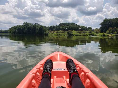 A person’s feet in sneakers resting on the edge of an orange kayak, reflecting clouds and greenery on calm water.
