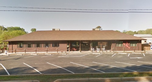 A single-story wooden building with a brown exterior, surrounded by a parking lot and trees in the background.
