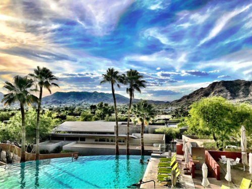 A scenic view of a pool surrounded by palm trees, mountains, and a vibrant sky with clouds.