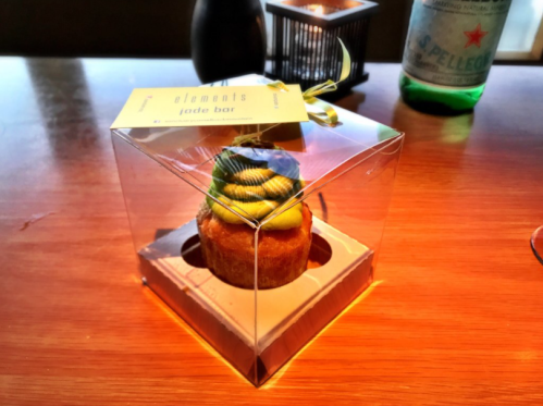 A cupcake in a clear box, topped with colorful frosting, sitting on a wooden table next to a bottle of sparkling water.