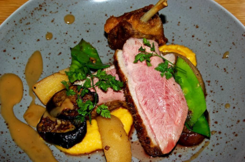 A gourmet dish featuring sliced pink duck breast, vegetables, and a sauce, elegantly arranged on a gray plate.