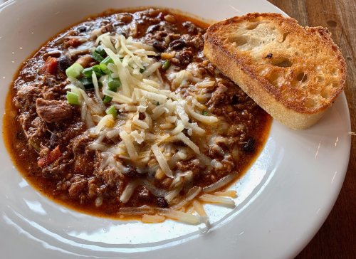 A bowl of hearty chili topped with cheese and green onions, served with a slice of toasted bread on the side.