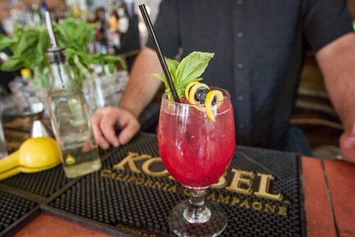 A vibrant cocktail garnished with basil and lemon twists, served in a glass on a bar counter.